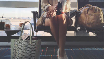Woman waiting at airport holding sunglasses and coffee with luggage on seat to her left and muffin on table to her right, with carry on bag on the floor below table